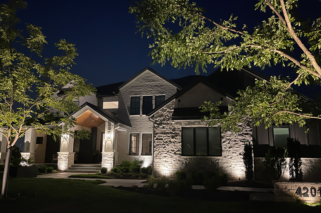 A large, two-story stone and brick house at night featuring professional architectural uplighting on the facade, illuminated entryway pillars, a lit address marker, and accent-lit landscaping trees.