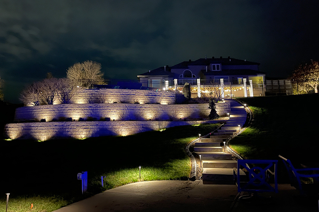 A wide nighttime view of a large home with three tiers of illuminated stone retaining walls and a winding concrete staircase featuring modern path lights leading up a grassy hill.