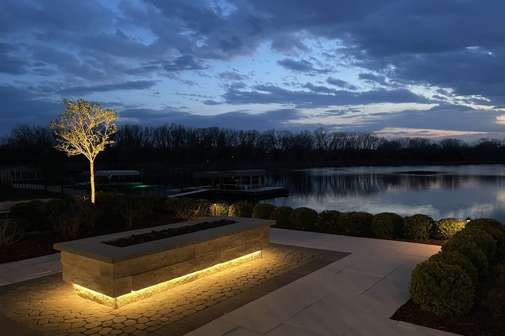A stone patio at twilight featuring a rectangular stone fire pit with glowing base lighting, an illuminated tree, and a scenic view of a lake with boat docks in the background.
