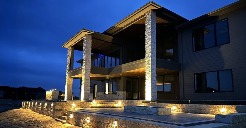 A nighttime view of a modern two-story home featuring tall stone pillars and a multi-level paver patio illuminated by landscape lighting in Wahoo, NE.