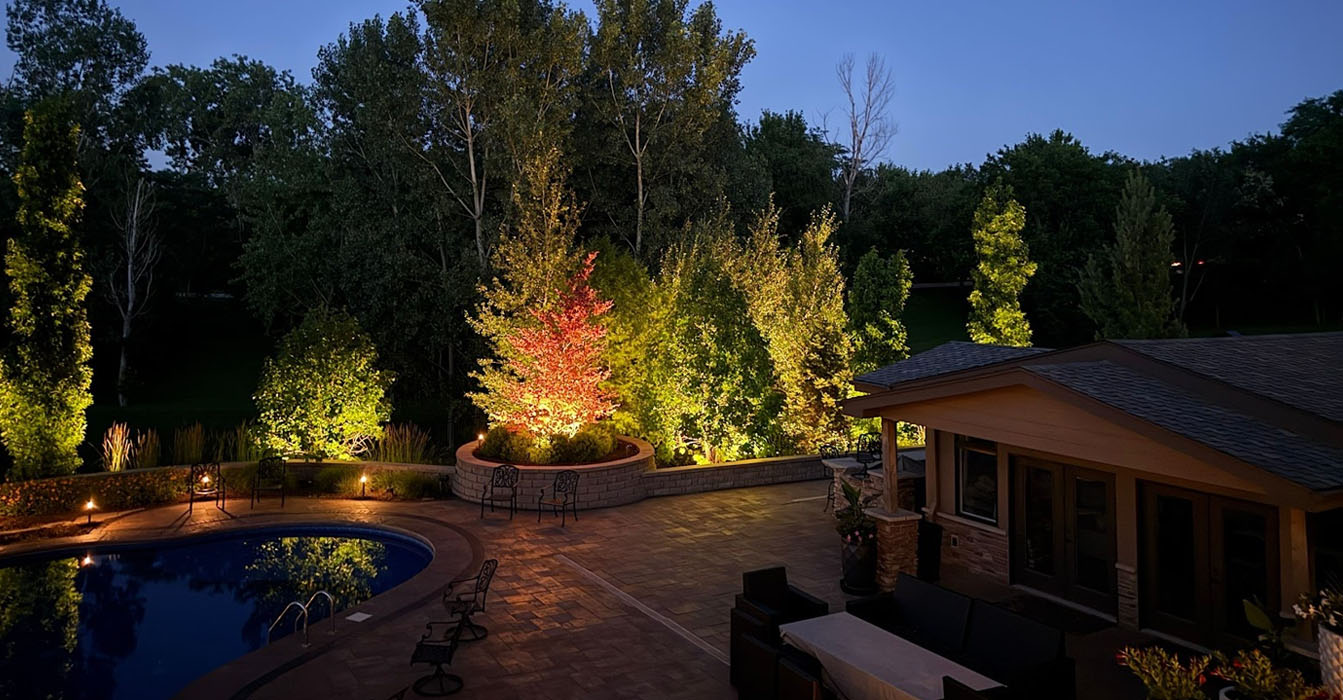 Backyard pool area illuminated with professional landscape lighting design showcasing trees and patio lighting at dusk.