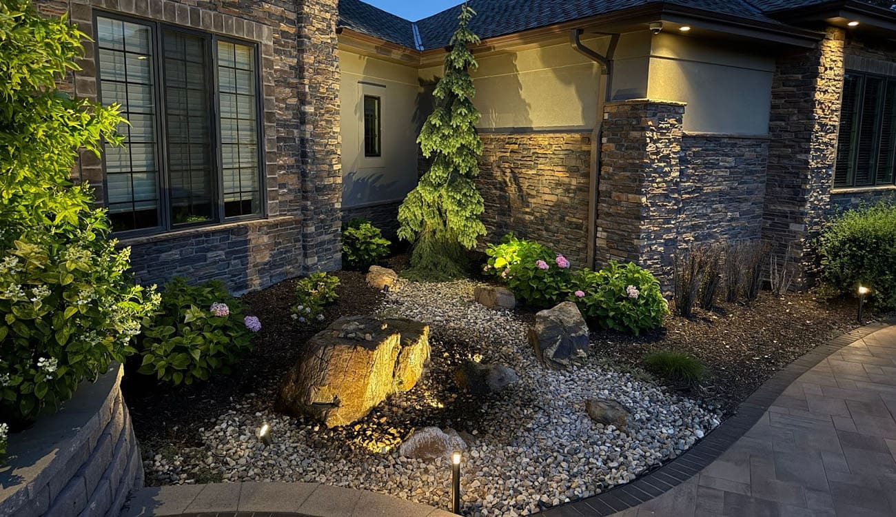 Front yard landscape lighting design featuring accent lights on stone walls, plants, and trees for a warm home exterior look.