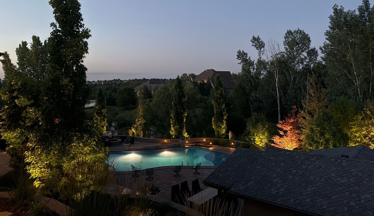 Golden lights reflect off a pristine pool, highlighting tall trees and a modern outdoor lounge.