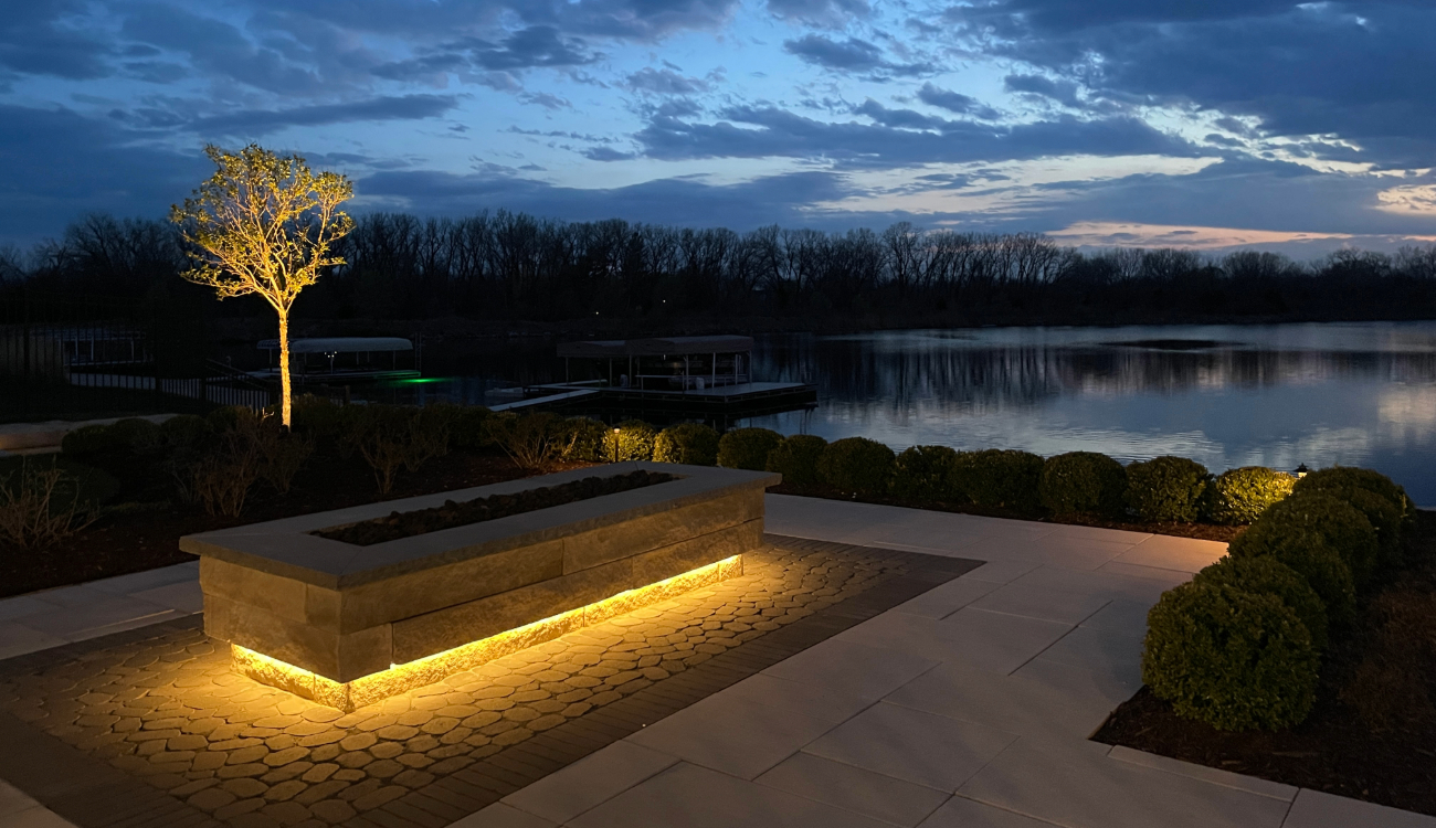 Stone fire pit and walkway glow with golden outdoor led lighting near the lake.