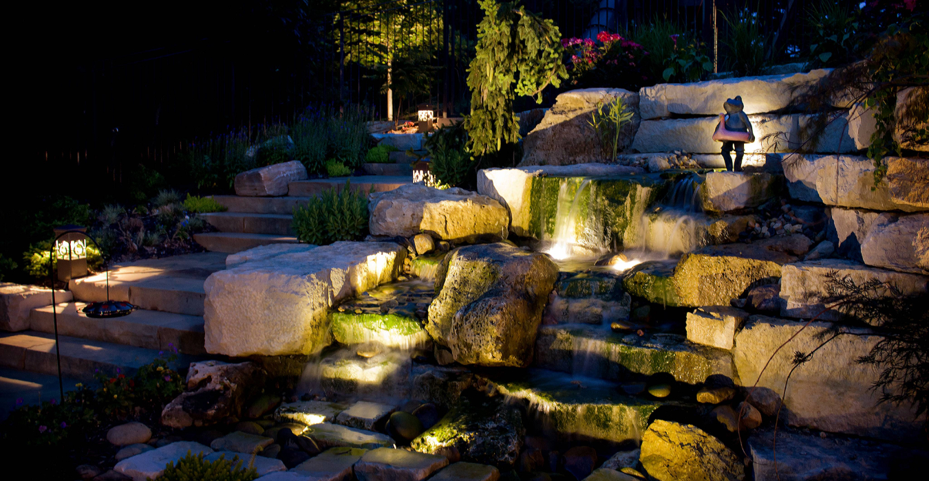 A backyard rock waterfall and stone steps illuminated at night, showcasing custom landscape lighting services.