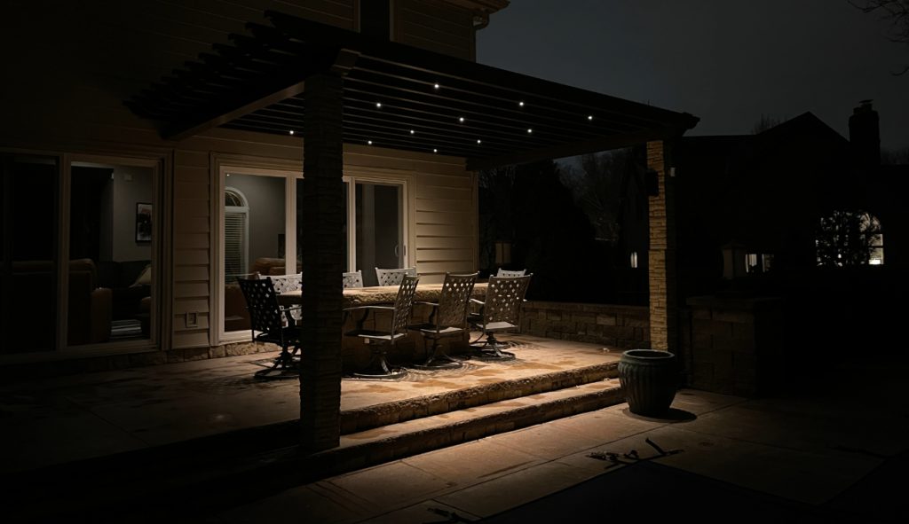 A nighttime view of a backyard patio pergola over a dining table, featuring small recessed patio lights.