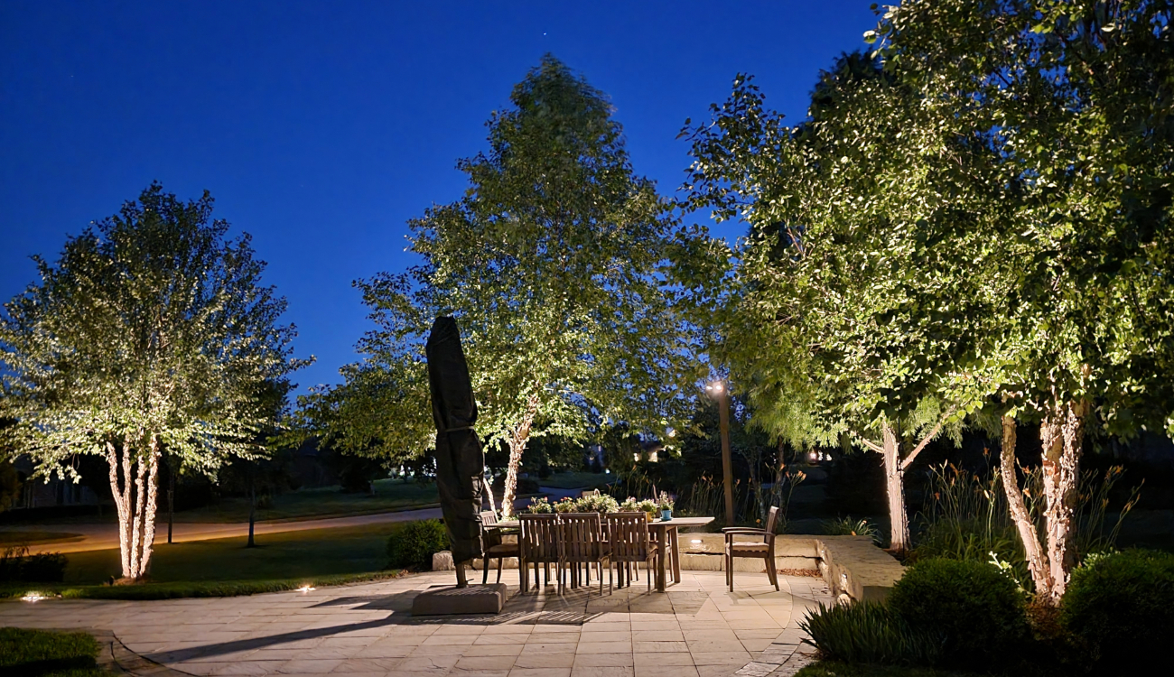 An outdoor stone patio with a dining table surrounded by trees illuminated by bright patio lights.